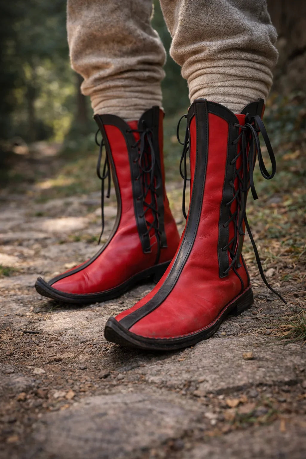 Vibrant red medieval leather boots with black laces, worn on a forest path for LARP or SCA reenactment.